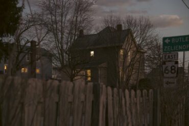 A dusk image of a house in a small town, Evans City, looking over a fence at a home with it's windows soft glow peaking out of the dark.